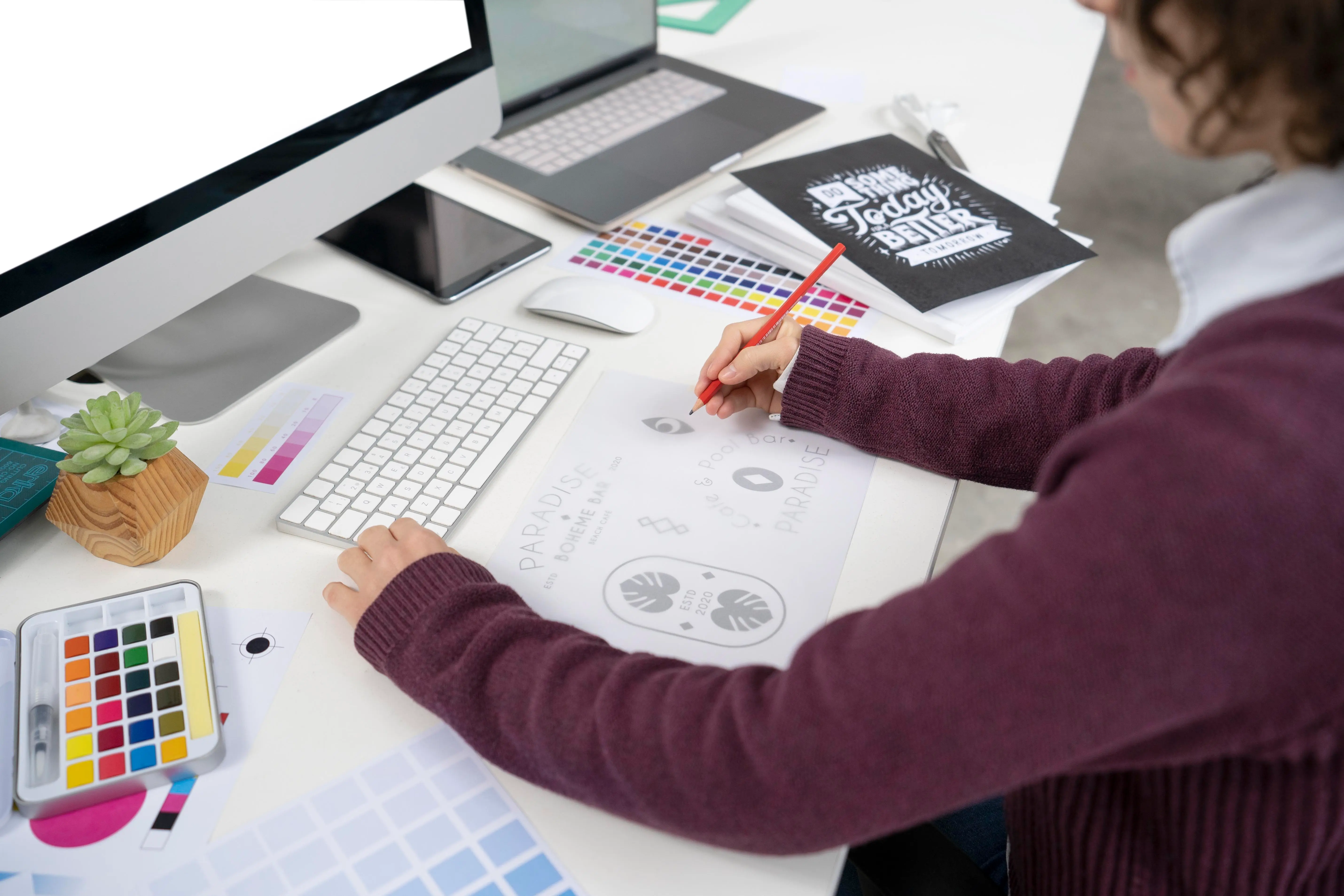 Person sketching graphic elements and the word "PARADISE" at a creative desk setup with design tools, computer, watercolor set, and motivational notebook—highlighting artistic workspace and design process.
