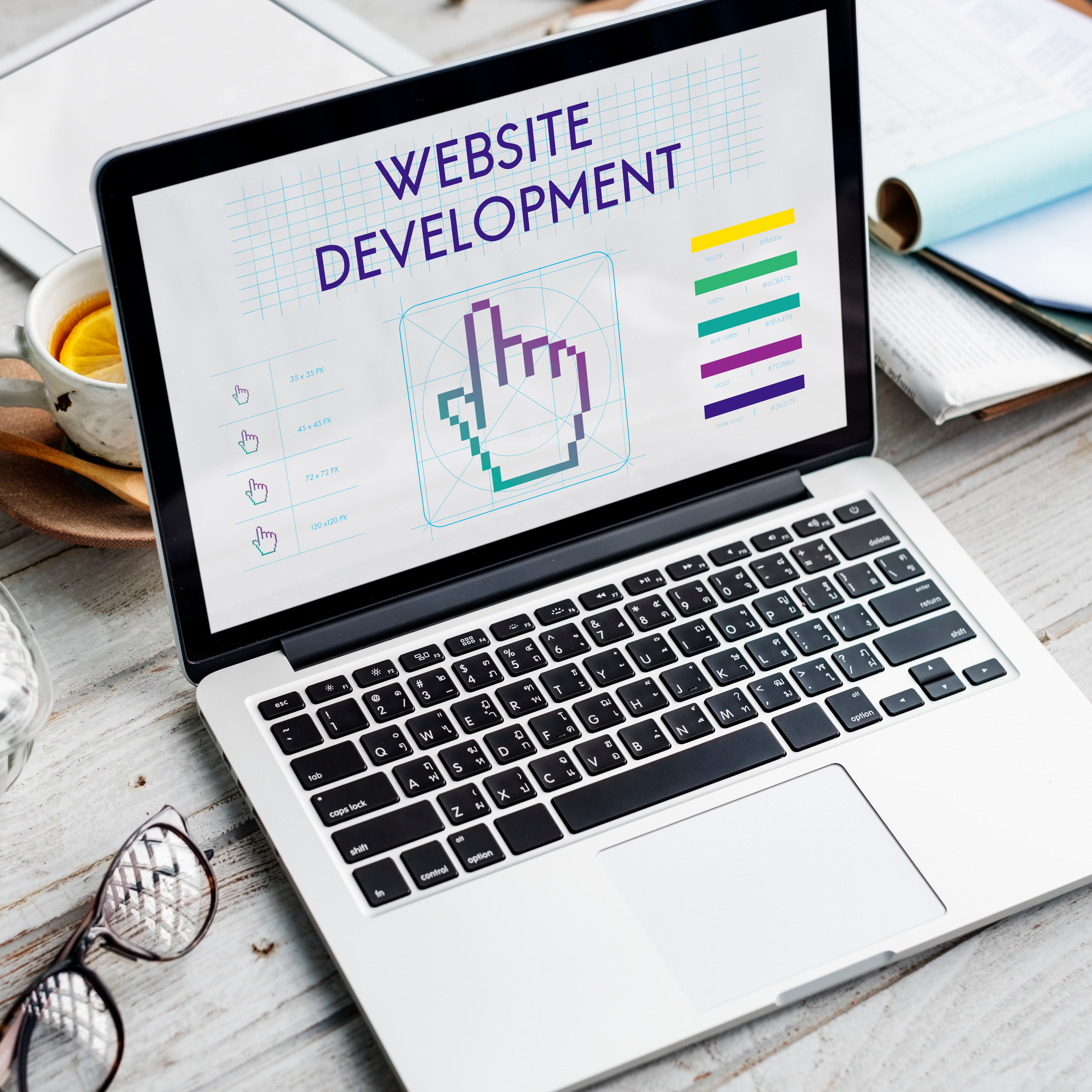 Laptop on wooden desk displaying a "Website Development" graphic, surrounded by workspace items like tea, glasses, notebook, and pen.