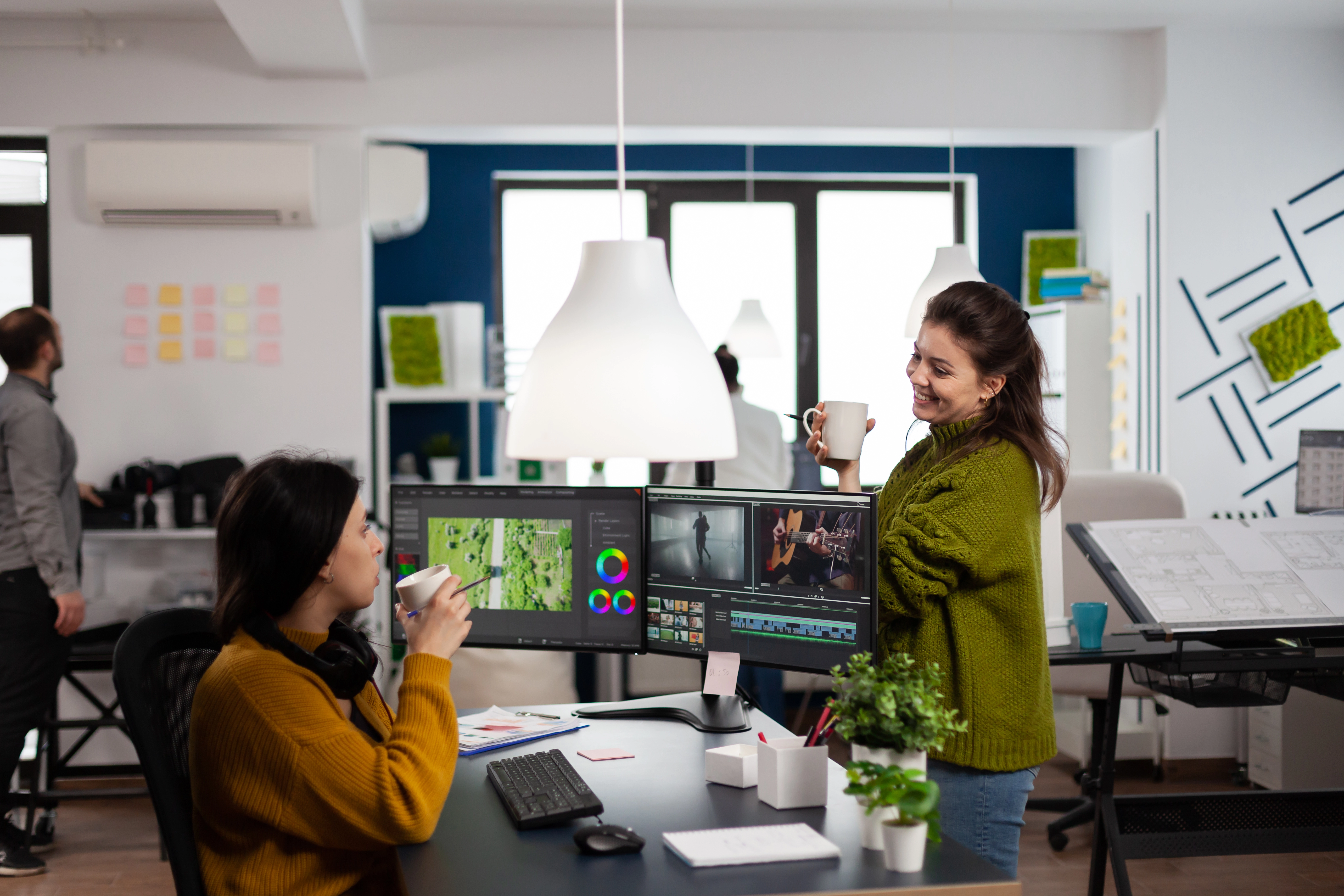 Two women in a modern office discussing video editing at a dual-monitor desk, possibly collaborating on an animation service project.