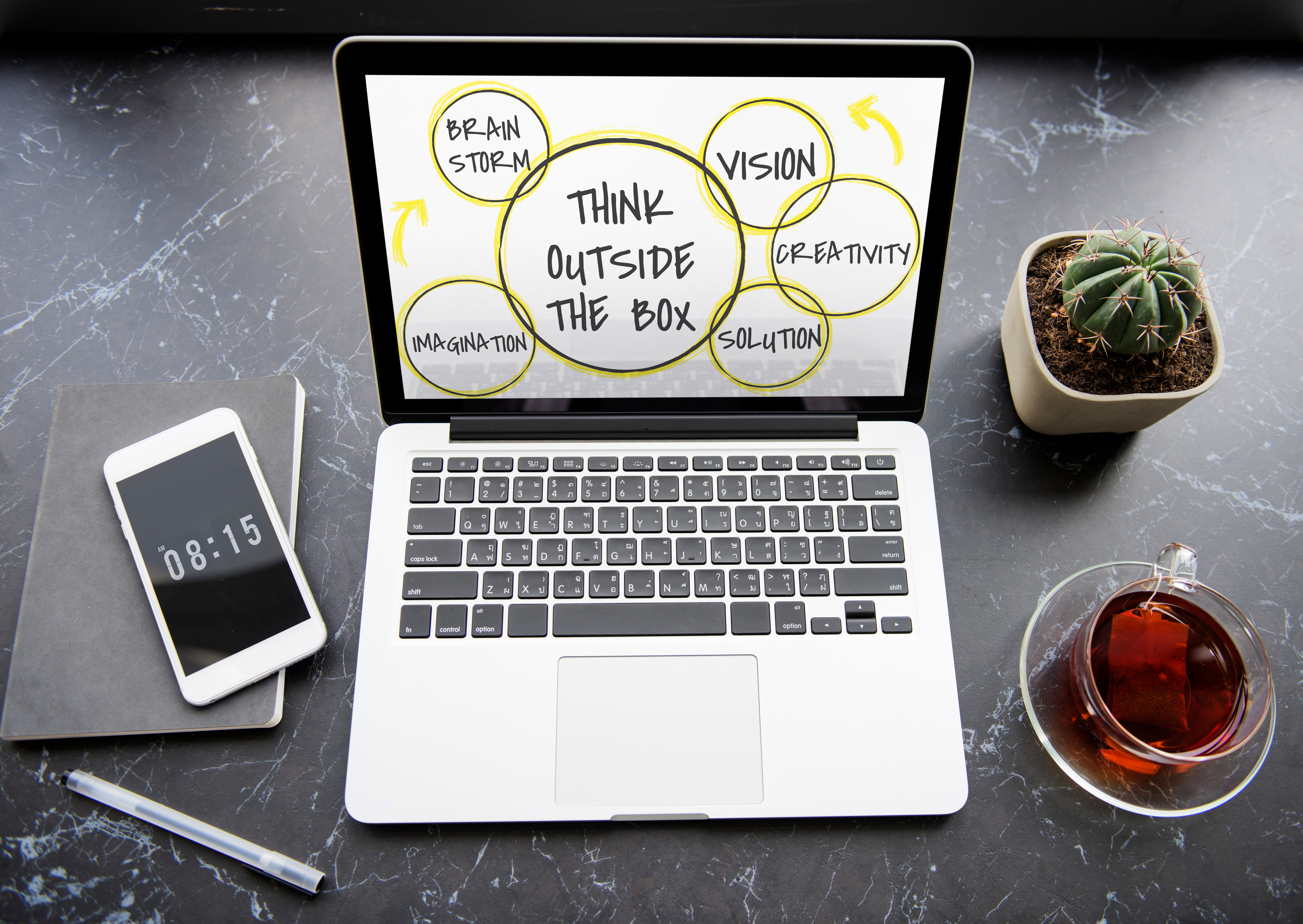 A laptop displaying a "Think Outside the Box" diagram with circles labeled "Brainstorm," "Imagination," "Vision," "Creativity," and "Solution," accompanied by a smartphone, notebook, pen, a cactus plant, and a cup of tea on a desk.